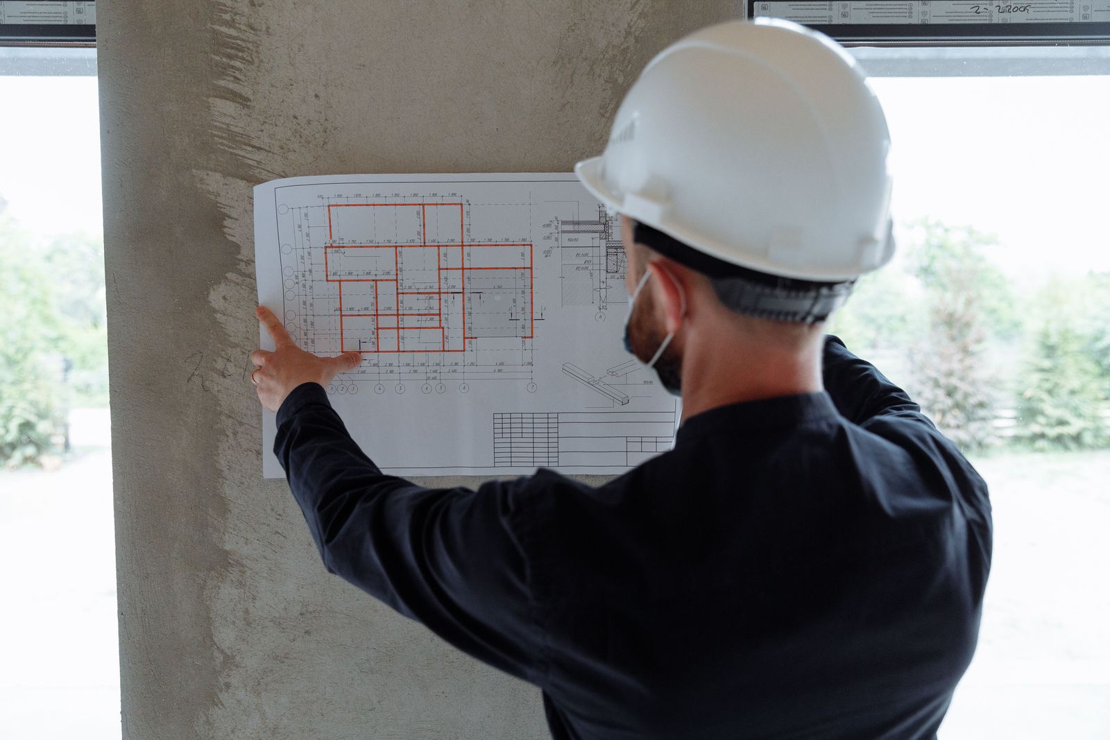 Person mit weißem Schutzhelm von hinten zeigt auf einen Bauplan, der an einer Betonsäule hängt (Symbolfoto)